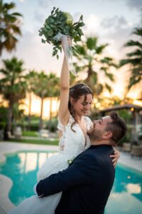 Lo sposo solleva in braccio la sposa davanti alla piscina al tramonto