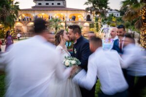 Bacio degli sposi circondato dagli invitati in movimento, villa illuminata sullo sfondo